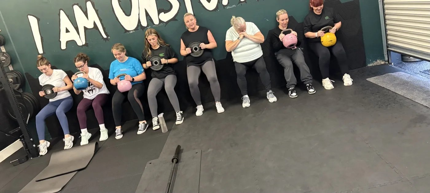 Lyndsay's group class lined up against a gym wall holding weights